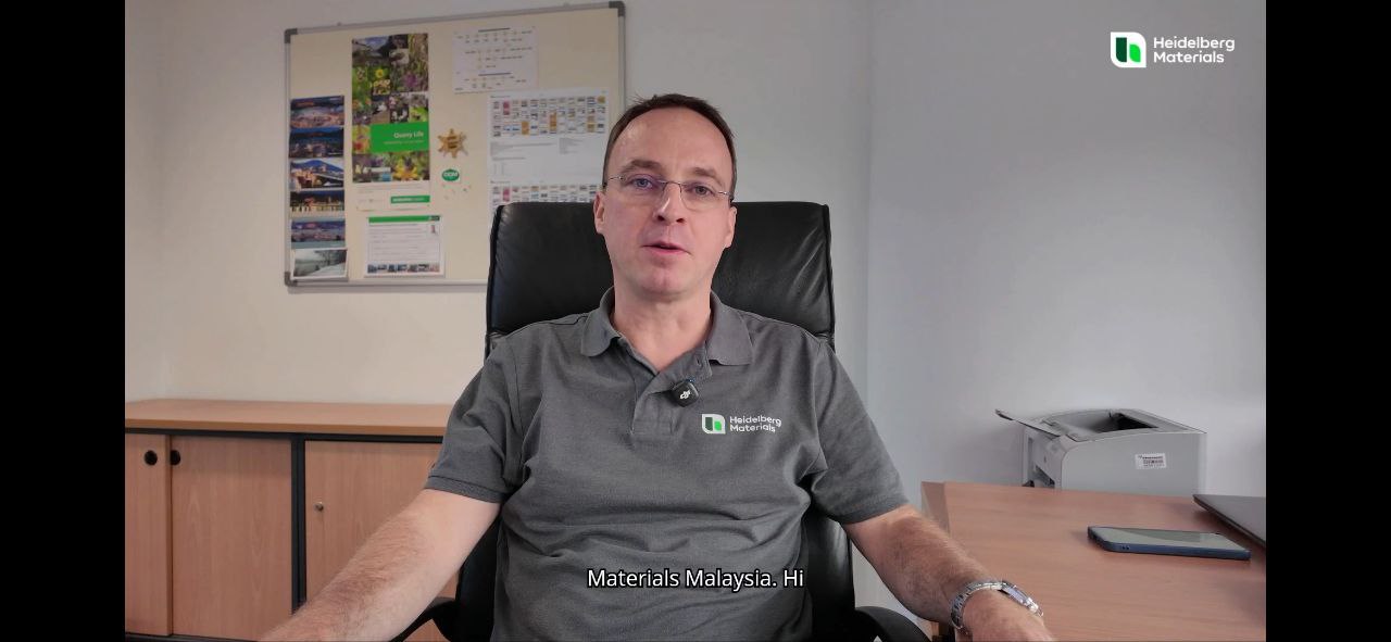 A person seated in an office chair wearing a gray polo shirt with a Heidelberg Materials logo on the chest.